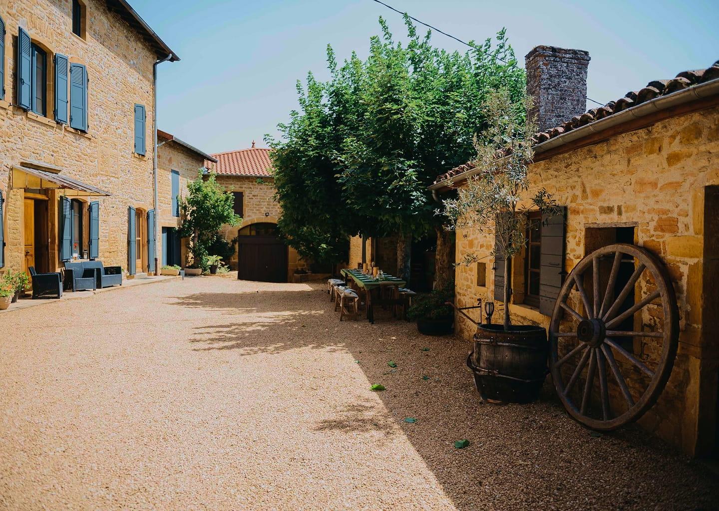 La cour en pierre dorée du domaine, avec ses tonneaux, sa roue de charrette et la grande table sous le platane